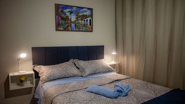 A neatly made bed with patterned bedding, two bedside lamps, a nightstand with a vase of flowers, and a painting of a street scene above the bed. - Apto com rea de Lazer a 15 min do Aeroporto (Manaus)