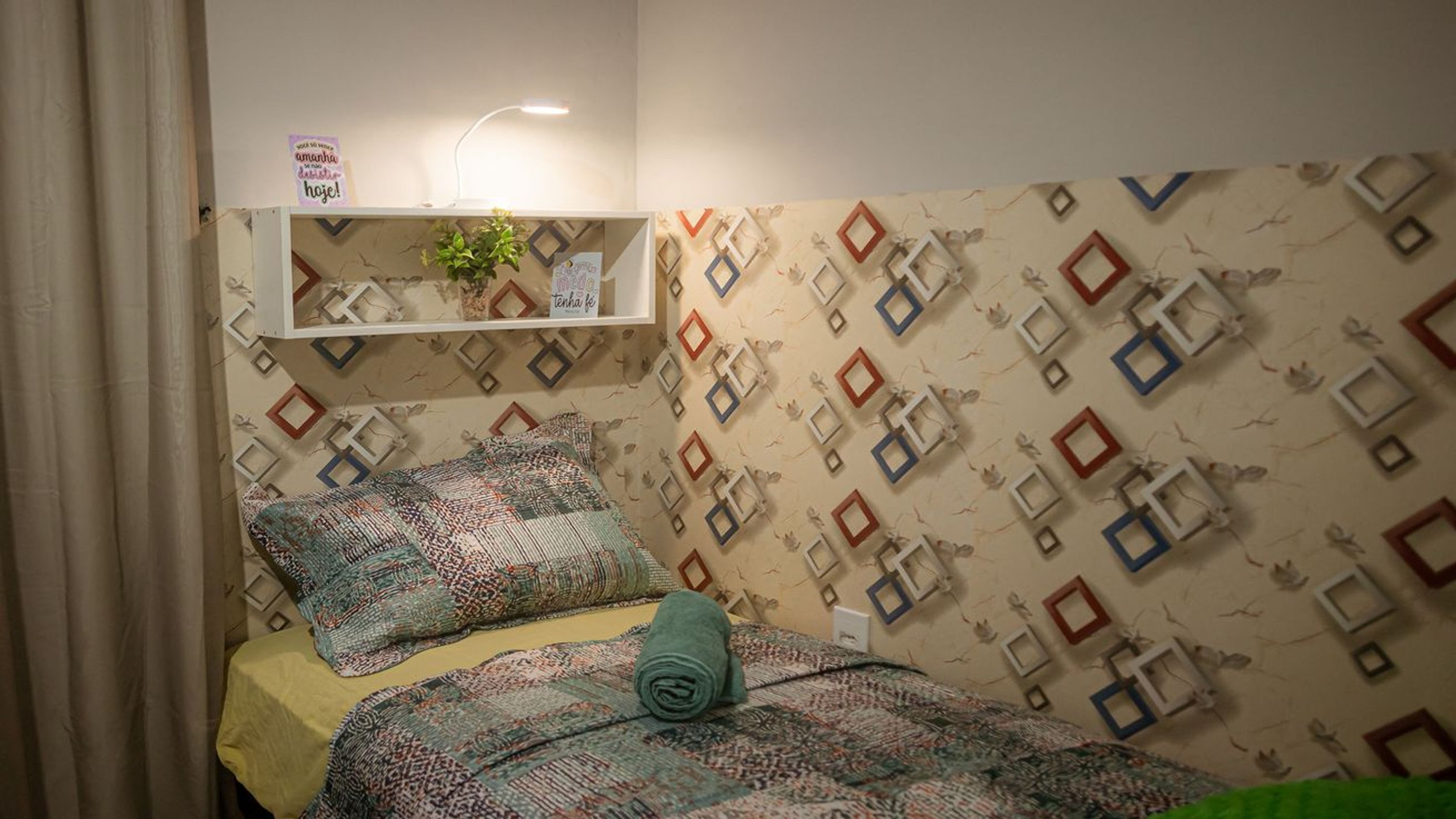 A bedroom with a patterned wall, a bed with a rolled-up blanket, a bedside table with a lamp, and a shelf with a plant and a sign.