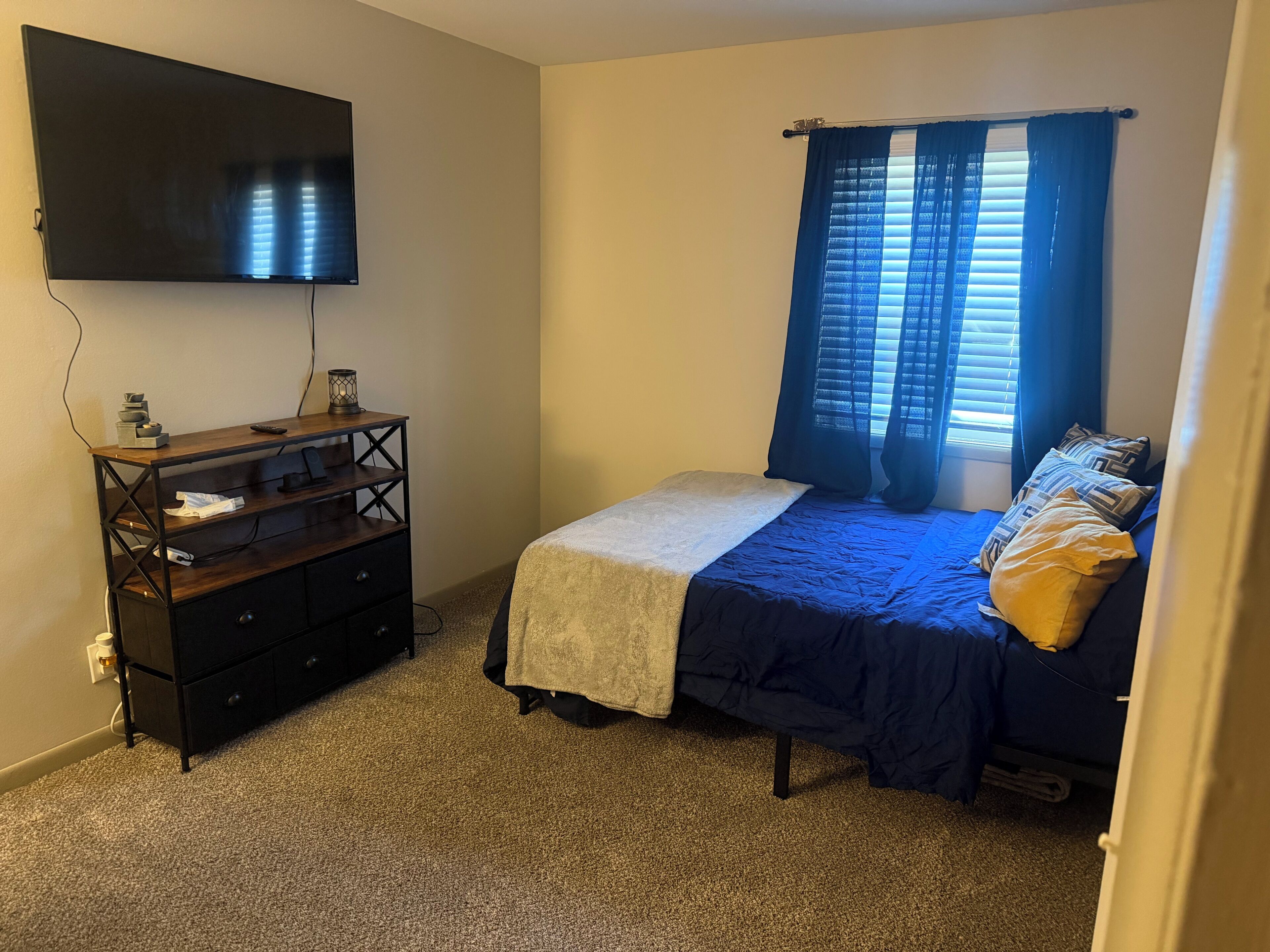 Une chambre à coucher avec un lit, un téléviseur fixé au mur, une commode et une fenêtre avec des rideaux.