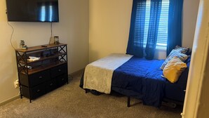 Un dormitorio con cama, televisor montado en la pared, cómoda y ventana con cortinas.
