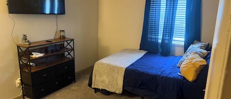 Une chambre à coucher avec un lit, un téléviseur fixé au mur, une commode et une fenêtre avec des rideaux.