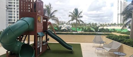 An outdoor play area with a slide and climbing structure, surrounded by palm trees and a view of a cityscape.