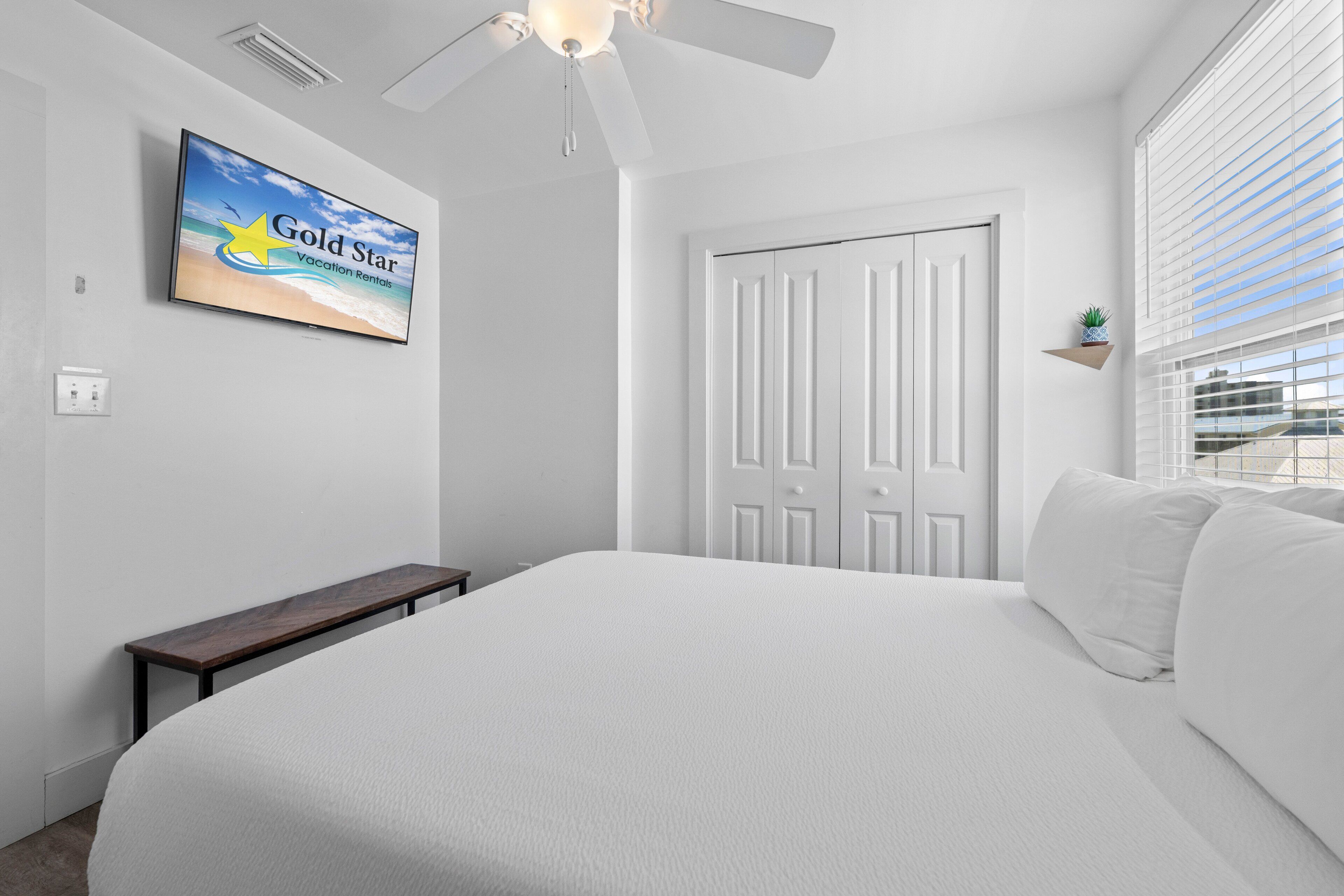 A bedroom with a bed, a ceiling fan, a TV on the wall, a door, and a window with blinds.