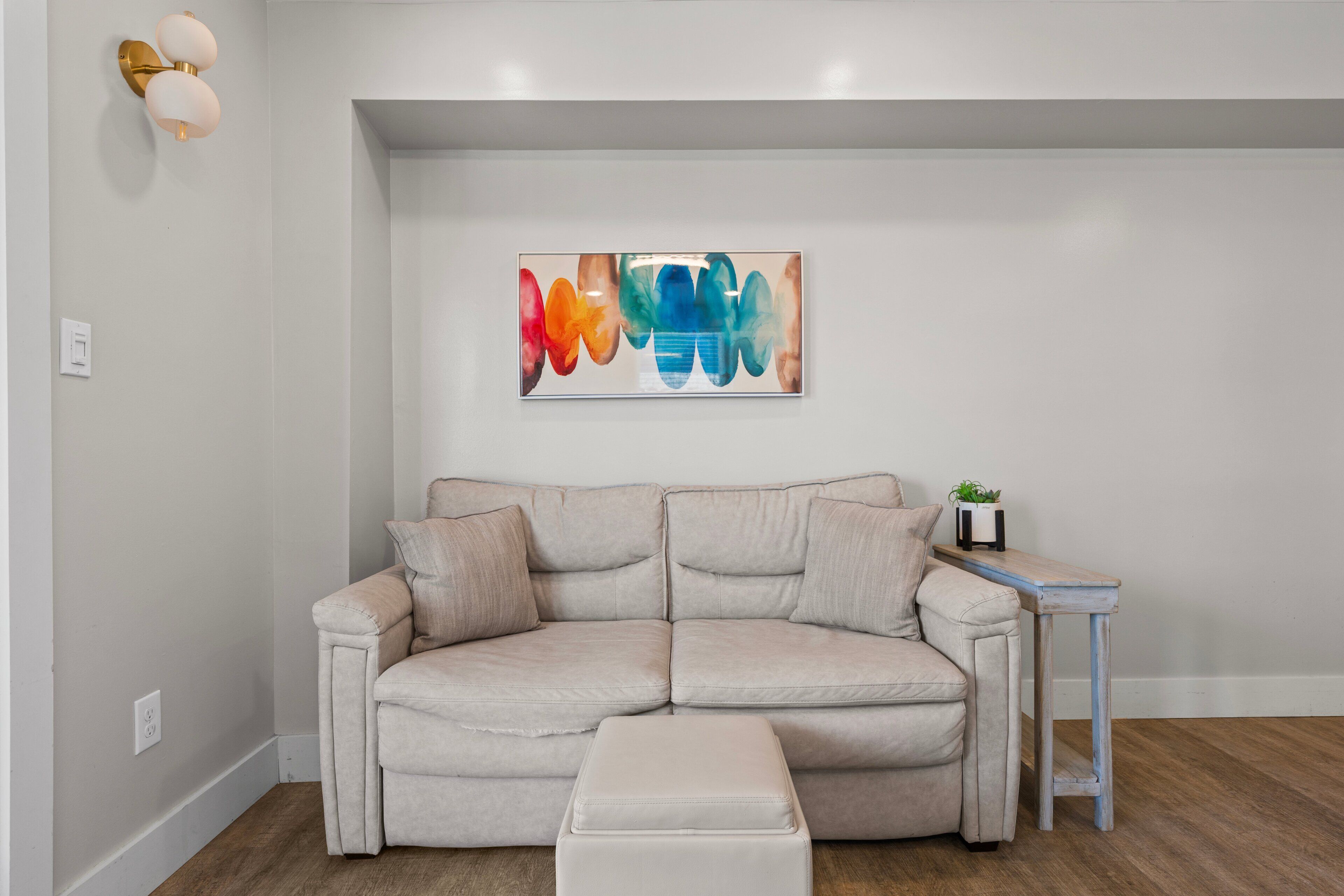 Ein sofasitz in neutralen Farbtönen mit passendem Hocker, ein kleiner Beistelltisch mit einer Pflanze und ein farbenfrohes abstraktes Gemälde an der Wand.