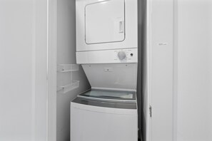 Ein kleiner, weißer Waschraum mit einer Frontlader-Waschmaschine und -Trockner, einer kleinen Ablage und einer Tür mit Beschriftung.