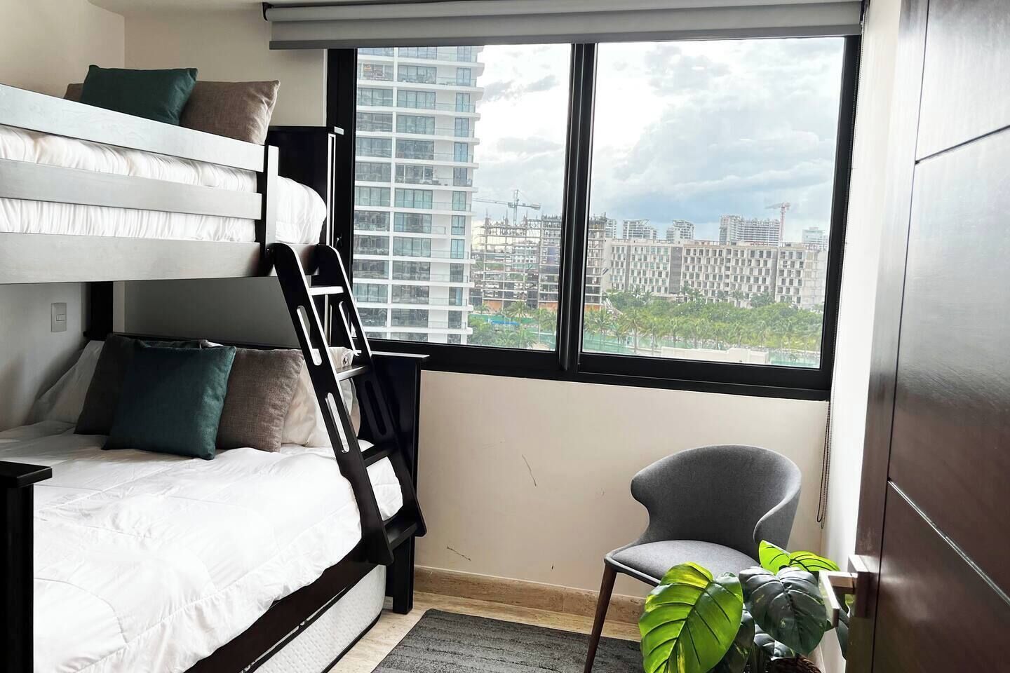 Une chambre avec un lit superposé, un bureau, une chaise, une fenêtre donnant sur des immeubles et une plante en pot.