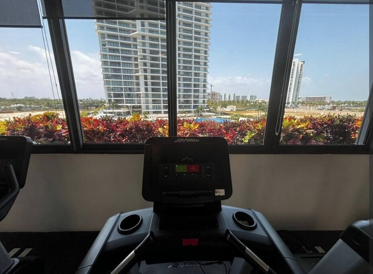 Una cinta de correr en una habitación con ventanas amplias que dan a un jardín cuidado y edificios altos.