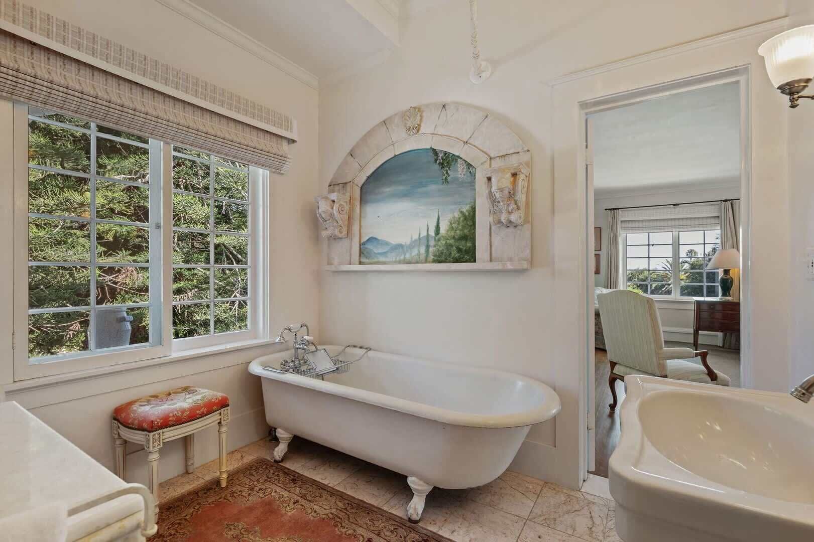 Ein Badezimmer mit freistehender Badewanne, einem Fenster mit Blick ins Grüne und einem Gemälde über der Wanne.
