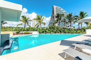 Una piscina con sillas de descanso, rodeada de palmeras y un edificio moderno al fondo.