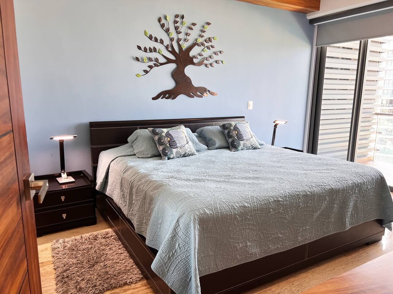 Ein Schlafzimmer mit einem Bett, Nachttischen, einer Wanddekoration mit Baummotiv und einem Fenster mit Jalousien.