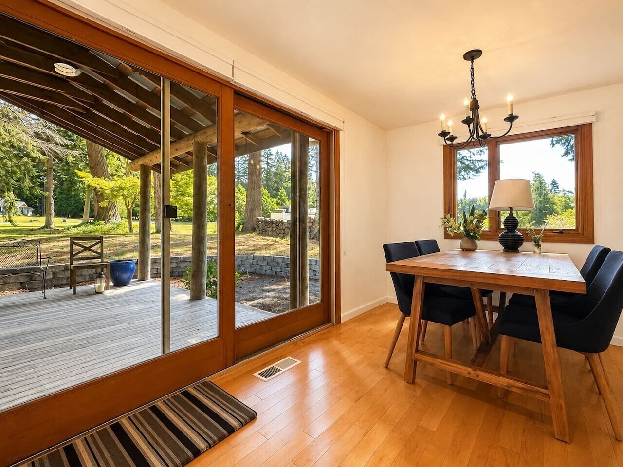 Zona pranzo con tavolo e sedie in legno, un lampadario e vista su un giardino tramite ampie porte scorrevoli in vetro.
