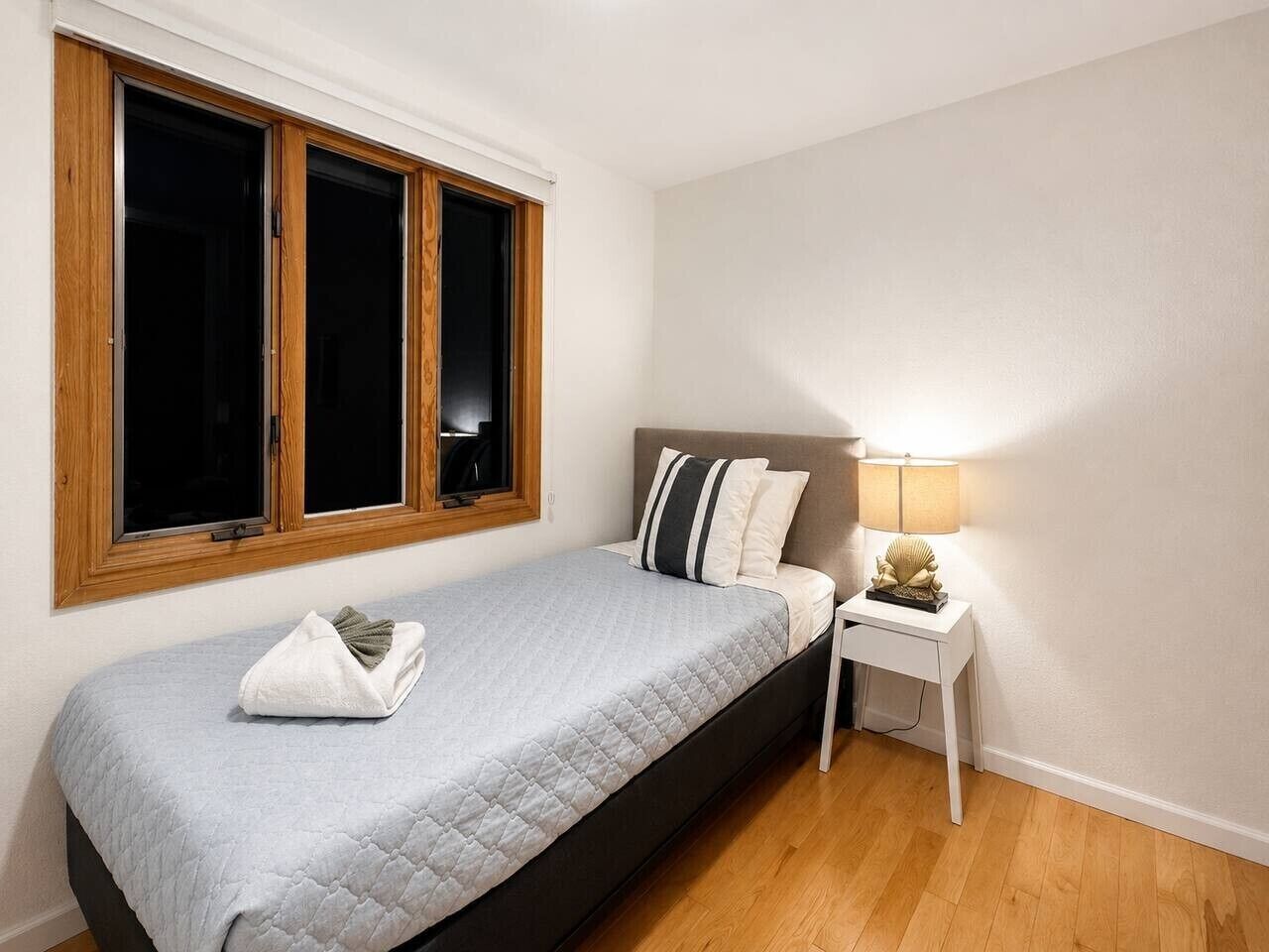 A single bed with a quilted cover, a bedside table with a lamp, and a window with wooden frames.