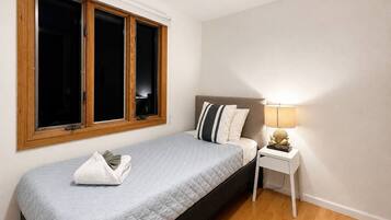 A single bed with a quilted cover, a bedside table with a lamp, and a window with wooden frames.