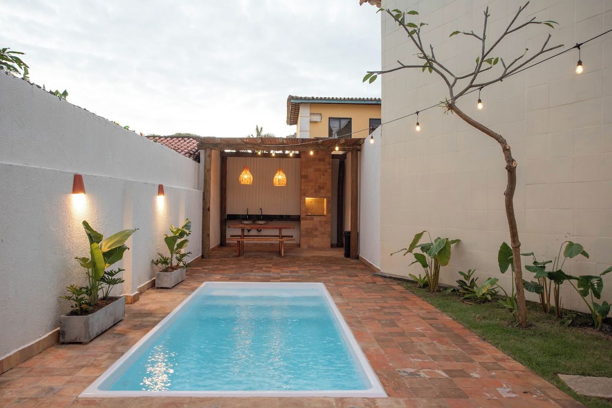 Piscina com deck revestido, área de churrasco e plantas em vasos.