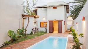 A swimming pool with a tiled deck, surrounded by a white building with a wooden door and a small window. - Mar Stays - House 22A (Salvador)