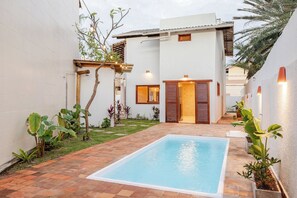 Une piscine avec une terrasse carrelée, entourée d’un bâtiment blanc doté d’une porte en bois et d’une petite fenêtre.