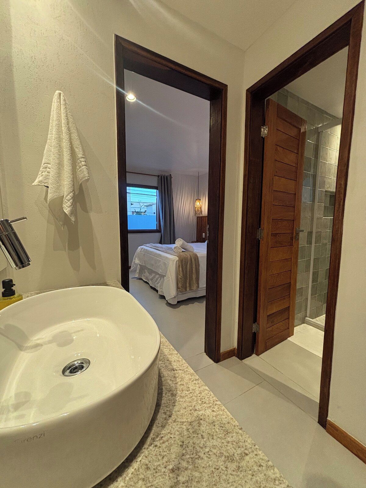 Une salle de bain moderne avec un grand lavabo, une douche et un lit visible à travers une porte ouverte.