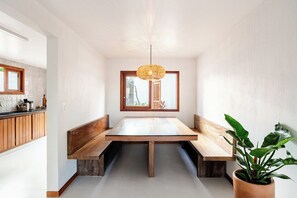 Een eetgedeelte met een lange tafel, banken, een hanglamp en een potplant.