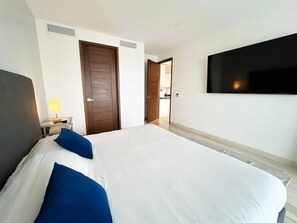 Quarto de hotel com cama, televisão na parede, porta e abajur em uma mesa de cabeceira.