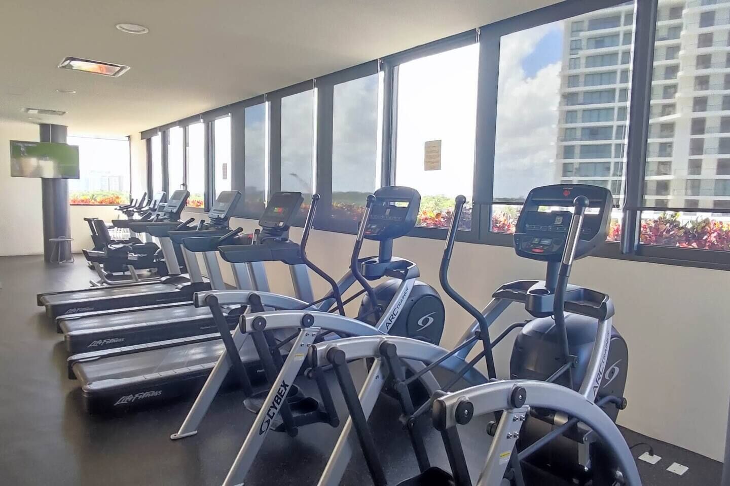 Ein Fitnessstudio mit Laufbändern und Ellipsentrainern, großen Fenstern und Blick auf ein Gebäude.