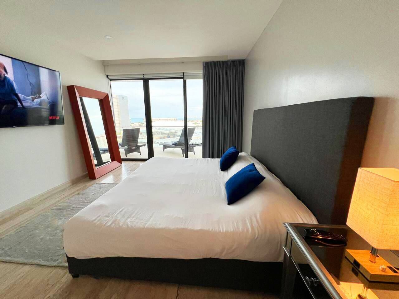 Ein Hotelzimmer mit einem großen Bett, einem an der Wand befestigten Fernseher, Blick ins Freie und einem kleinen Tisch mit Lampe.
