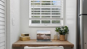 Ein Laptop auf einem Holztisch, darüber ein Fenster, eine Topfpflanze und ein kleiner Tisch mit einer Kerze.