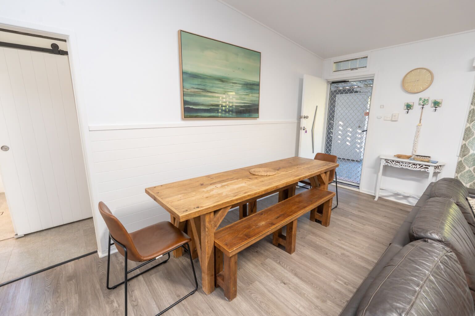 A dining area with a wooden table and benches, a sofa, and a painting on the wall.