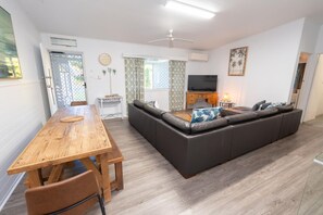 A modern living room with a wooden dining table, a sofa set, a television, and a ceiling fan. - A Little Grey Shack - Beach Cottage Amity Point (Amity Point)
