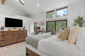A bedroom with a large bed, a TV, a dresser, and a desk with a chair. There are windows with a view of trees and a balcony with a grill. - Red Mountain Shadows Retreat: Sleeps 6 in West Sedona Near Hiking Trails, Park & Bike Paths (Sedona)