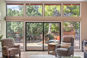 A spacious living room with large windows, a sliding glass door leading to a patio, and a view of a backyard with trees and a wooden fence. - Red Mountain Shadows Retreat: Sleeps 6 in West Sedona Near Hiking Trails, Park & Bike Paths (Sedona)