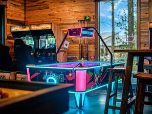 A neon-lit arcade table with a racing game interface, surrounded by wooden walls and a view of the outdoors. - Heated Pool + Pickleball | Pets | Sauna | Sleep 32 (Broken Bow)