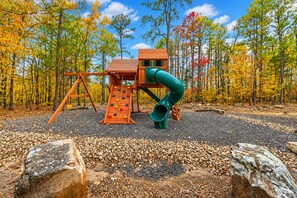 A playground with a slide and climbing wall, surrounded by trees with autumn leaves.