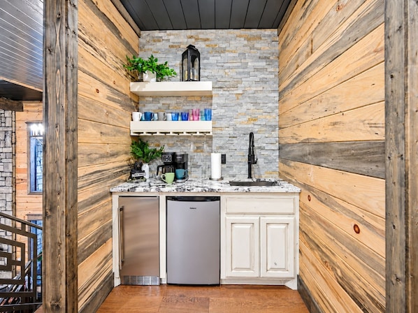 Wet bar with a sink, coffee maker, beverage refrigerators, open shelving, stone backsplash, and custom cabinetry.