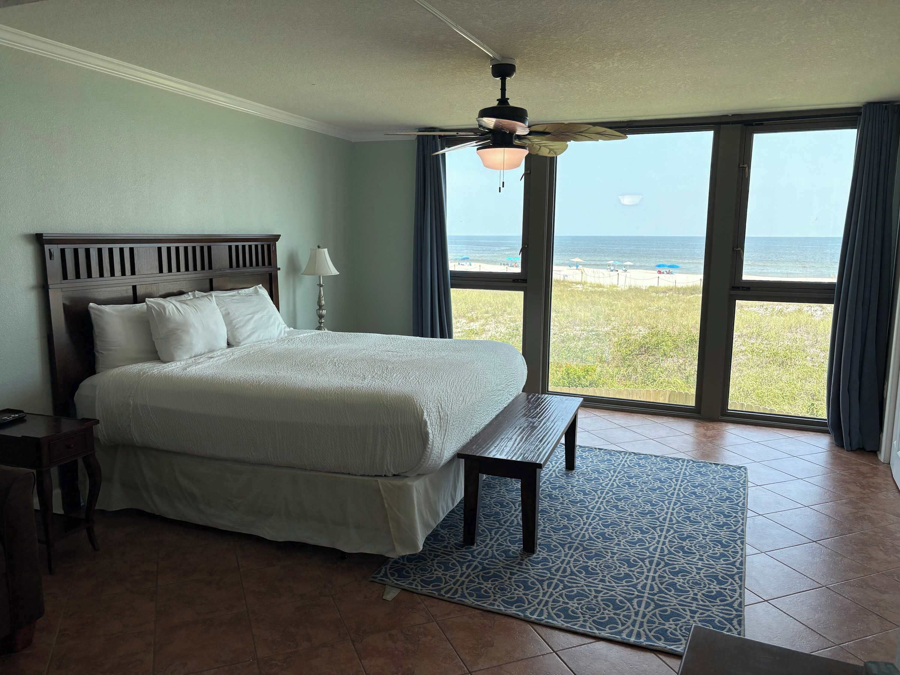 Un dormitorio con una cama grande, ventilador de techo, vista a la playa y piso de baldosas.