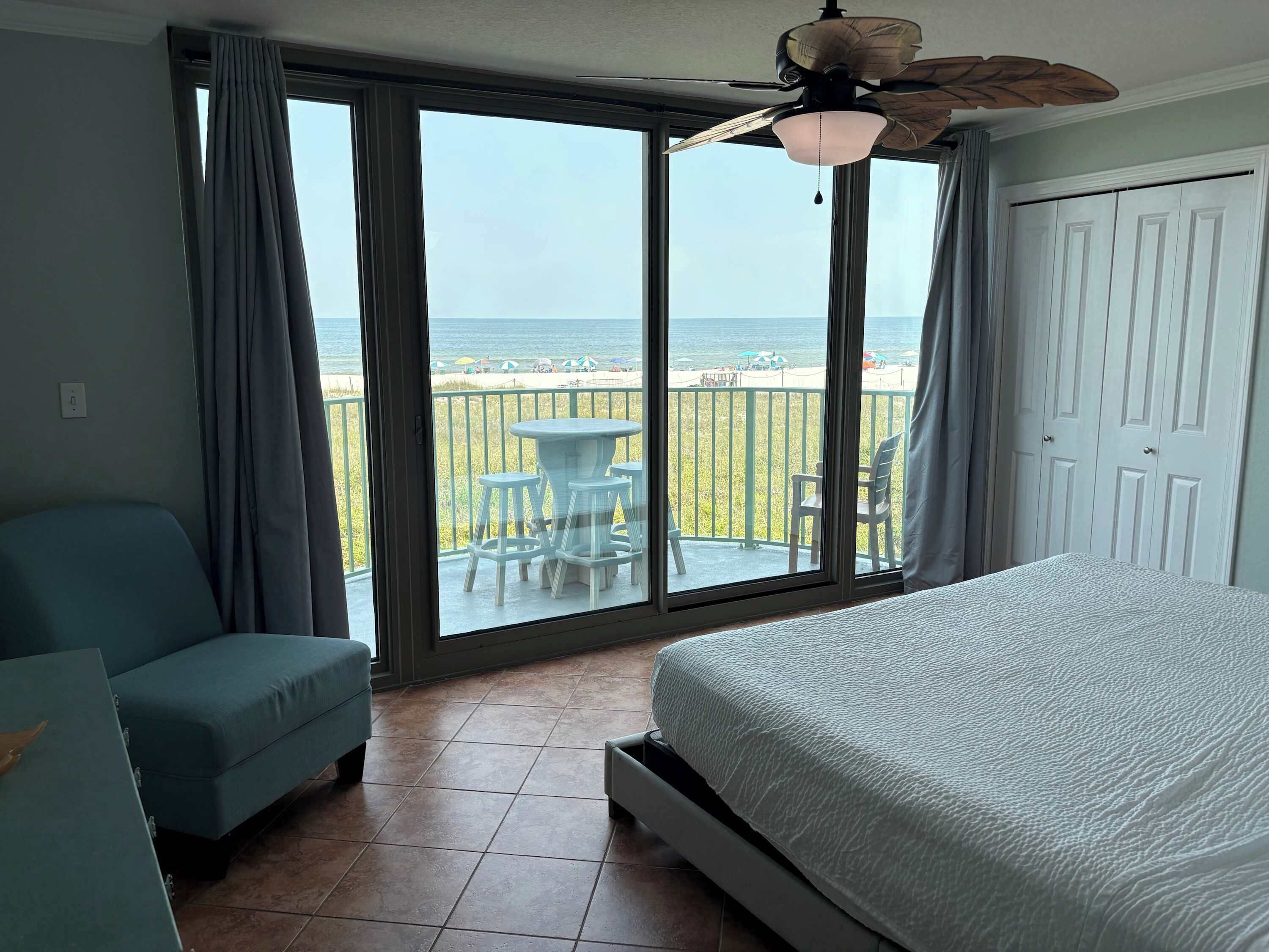 A bedroom with a bed, a chair, a table, and a view of the beach through large windows.