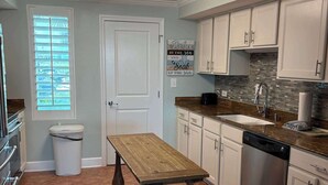 Una cocina con armarios blancos, una mesa de madera, lavavajillas y una ventana con persianas.