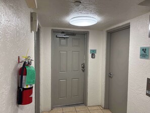 A hallway with a fire extinguisher mounted on the wall, a door with a number sign, and a light fixture on the ceiling. - Perdido Key Paradise Beachfront 3BR (Pensacola)