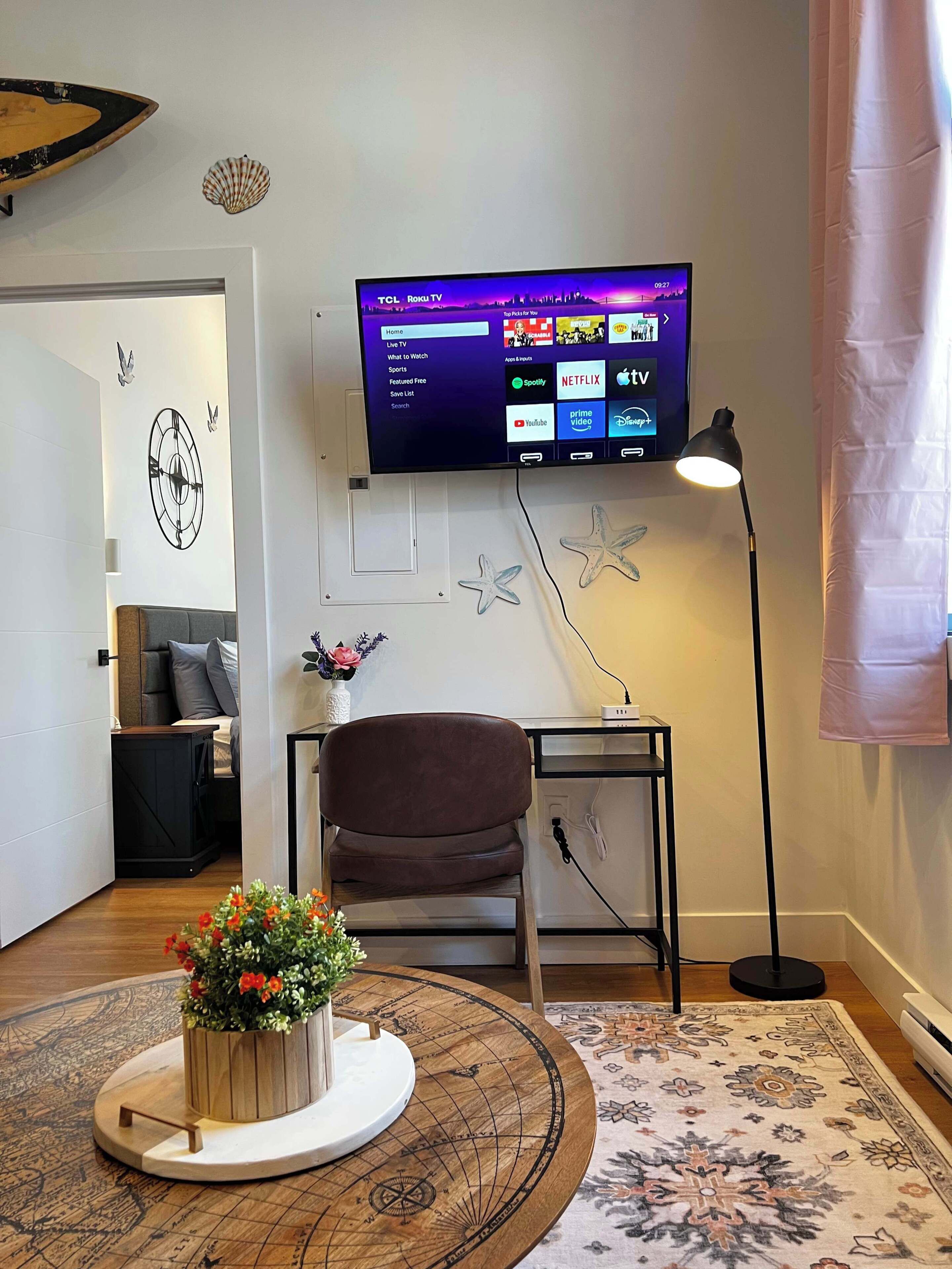 Una sala de estar moderna con un televisor de pantalla plana montado en la pared, un sillón de cuero marrón, una mesita auxiliar, una estrella de mar decorativa, una mesa redonda de madera con un arreglo floral, y una alfombra estampada.