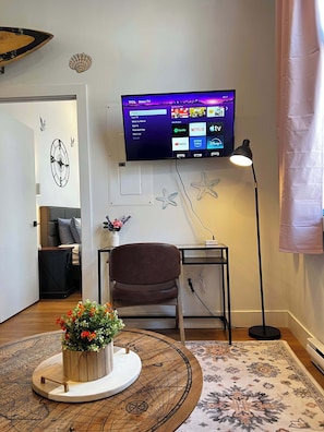 A modern living room with a flat-screen TV mounted on the wall, a brown leather chair, a small side table, a decorative starfish, a round wooden table with a flower arrangement, and a patterned rug. - Sunrise Surf Suite, Downtown Halifax (Halifax)