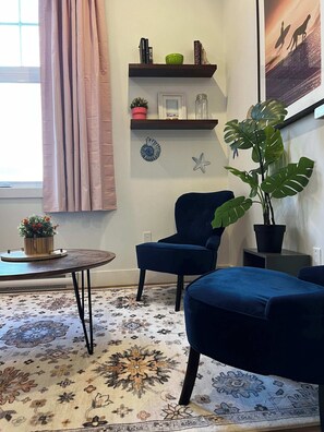 A living room with a blue armchair, a round coffee table, a potted plant, a bookshelf, and a decorative wall piece. - Sunrise Surf Suite, Downtown Halifax (Halifax)