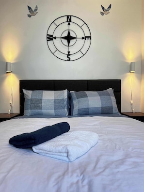 A neatly made bed with a folded towel and a pillow, a compass wall decoration, and two wall-mounted lamps. - Sunrise Surf Suite, Downtown Halifax (Halifax)