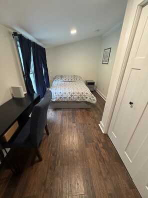 A bedroom with a bed, a desk, a chair, a bedside table, a small bedside lamp, and a door.