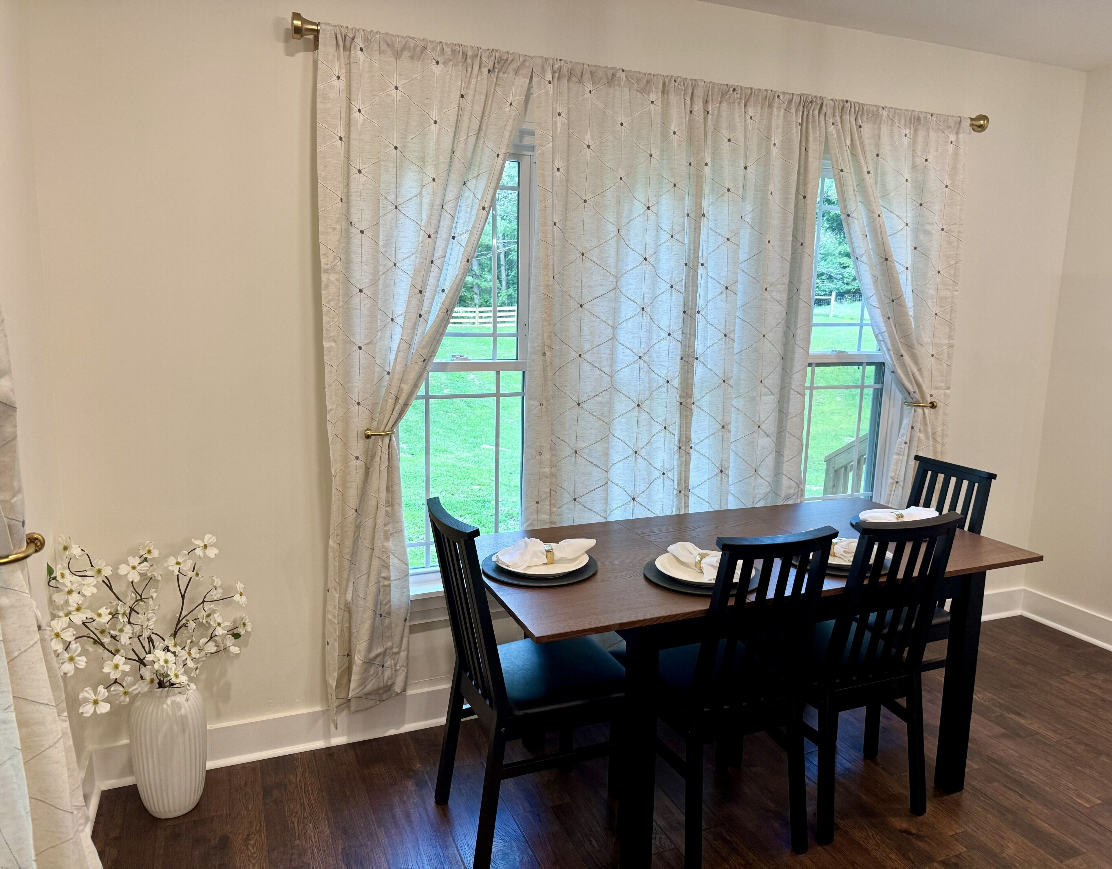 A dining area with a table set for four, a vase with white flowers, and a view of a green lawn through the window.