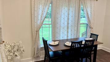 A dining area with a table set for four, a vase with white flowers, and a view of a green lawn through the window.