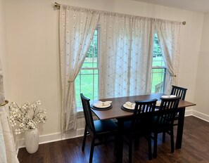 A dining area with a table set for four, a vase with white flowers, and a view of a green lawn through the window.
