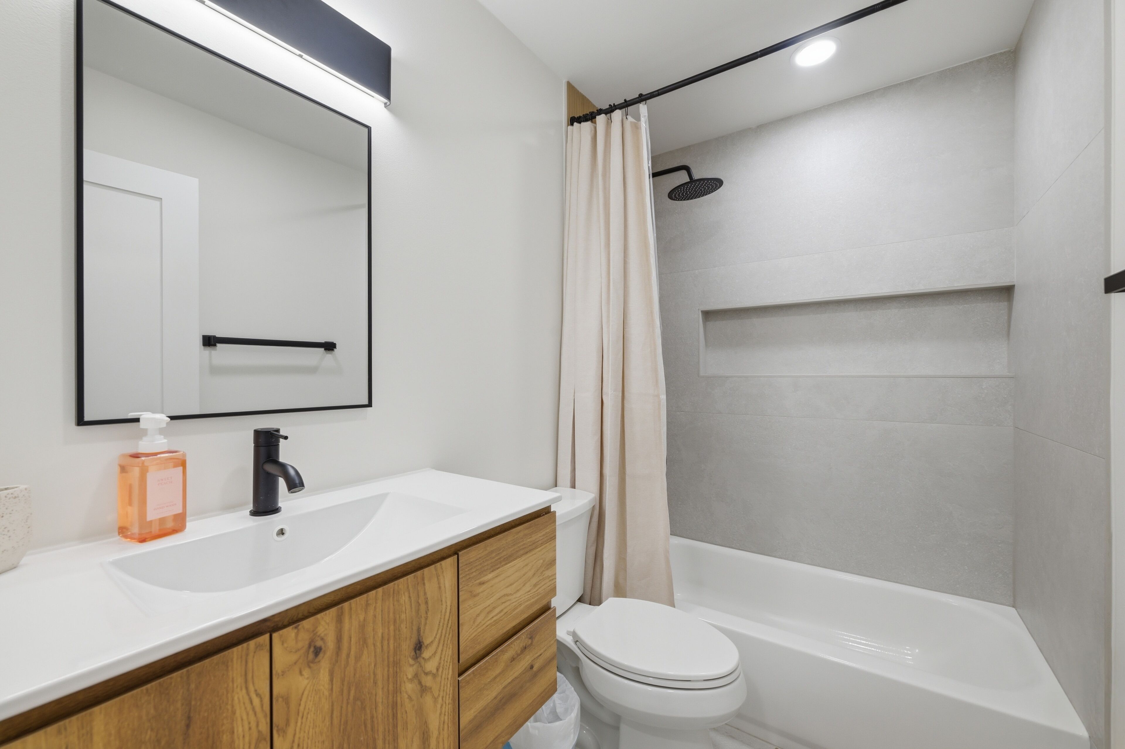 Un baño moderno con lavamanos blanco, tocador de madera, espejo, inodoro y zona de ducha.