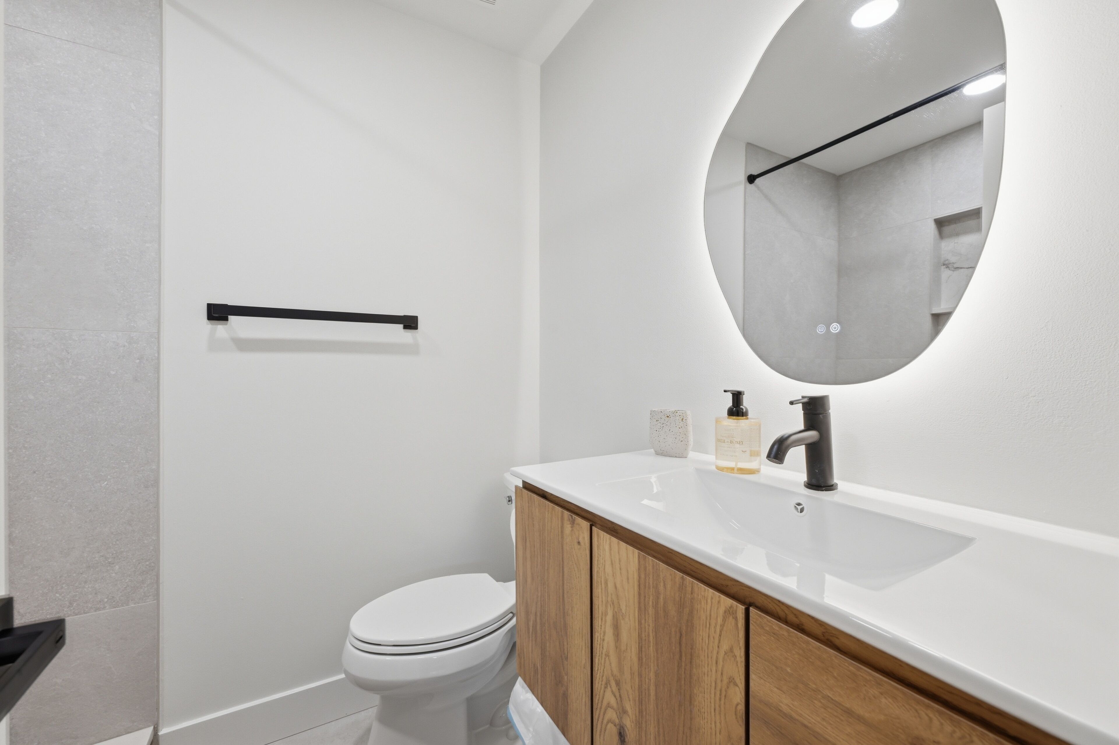 Une salle de bain moderne avec des toilettes blanches, un meuble-lavabo en bois, un miroir rond et un porte-serviettes noir.