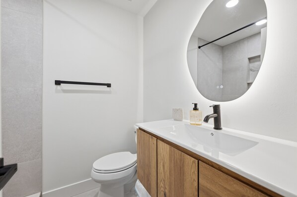 A modern bathroom with a white toilet, a wooden vanity, a round mirror, and a black towel rack.