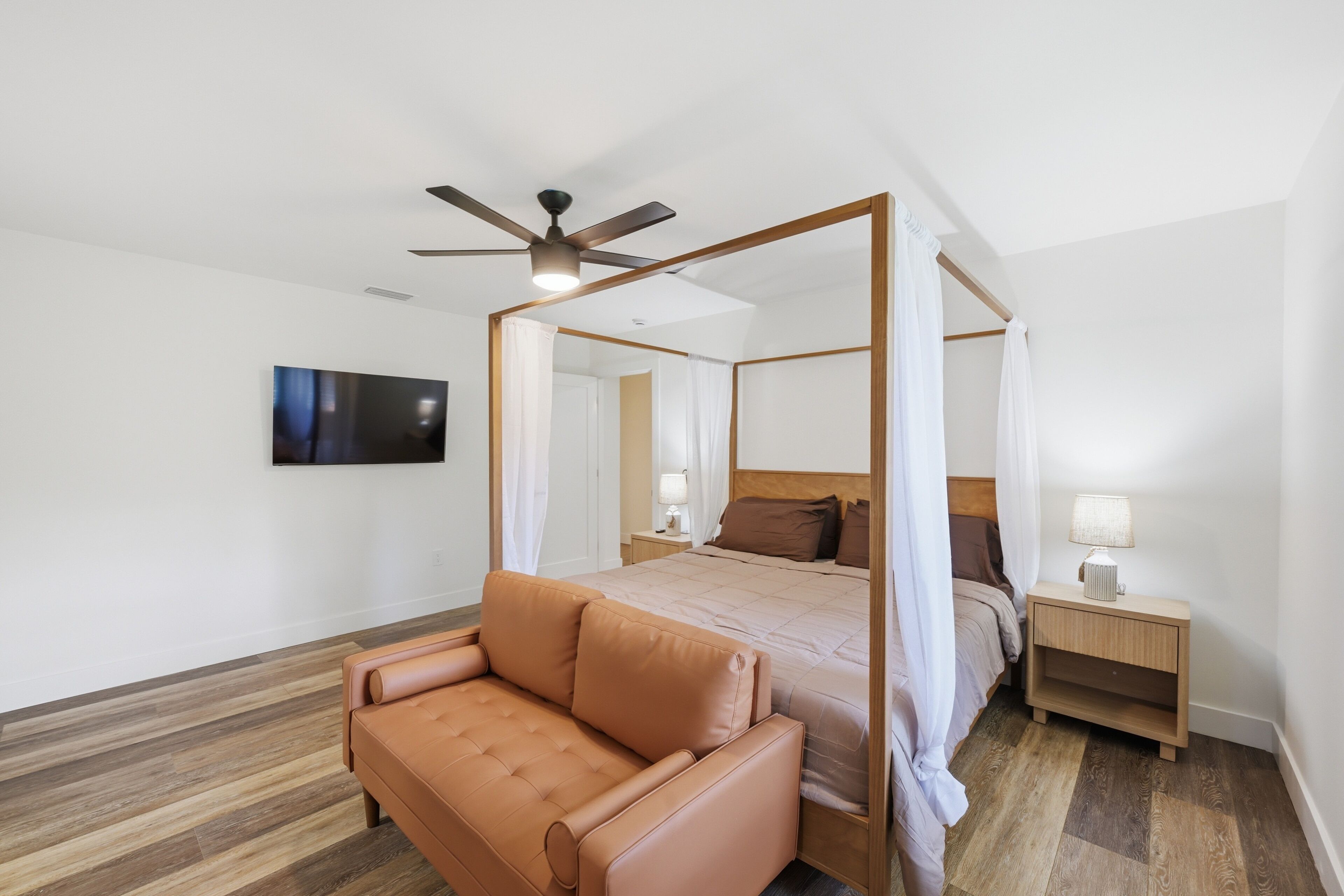 Un lit à baldaquin à quatre poteaux avec un ventilateur de plafond, un téléviseur à écran plat fixé au mur, une table de chevet en bois avec une lampe et un canapé en cuir.