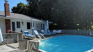 En swimmingpool med træterrasse, liggestole og en murstens skorsten.